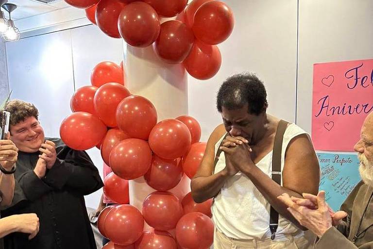 Homem aparece compungido, com as mãos em prece perto do rosto, emocionado. Ao fundo, balões de gás e cartazes mostram se tratar de uma festa de aniversário