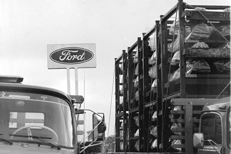 Caminhão com preças de veículos parado em pátio da Ford durante greve nos anos 1980, em frente a grande logotipo com o nome Ford escrito dentro de uma elipse escura