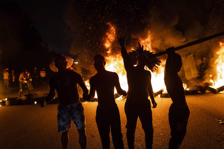 Quatro pessoas estão em pé, sem camisa, diante de fogo ateado a pneus
