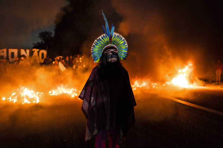 Indígena com cocar está em pé em rodovia. Atrás, há cartazes de protesto contra o marco temporal e fogo