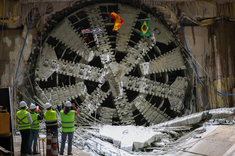 A imagem mostra uma grande máquina de perfuração de túnel, com uma roda de corte visível na parte frontal. Ao fundo, há bandeiras da Espanha e do Brasil penduradas. Na frente da máquina, um grupo de trabalhadores, usando coletes verdes, observa o equipamento. Fios e estruturas de suporte estão visíveis ao redor.