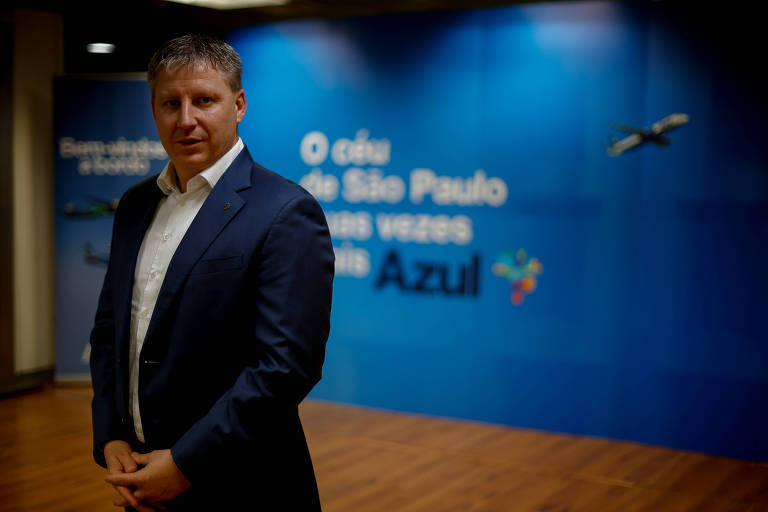 CEO da Azul posando para a foto com um banner da companhia aérea atrás. Ele está em pé, com os braços estendidos e as mãos entrelaçadas, rente ao seu corpo.