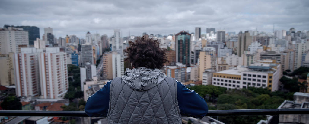 Ciro Schunemann é um homem negro de cabelos crespos. Ele está de costas, com agasalhos e olhando para o horizonte, com uma vista panorâmica de alguns prédios que compõem a cidade de São Paulo.