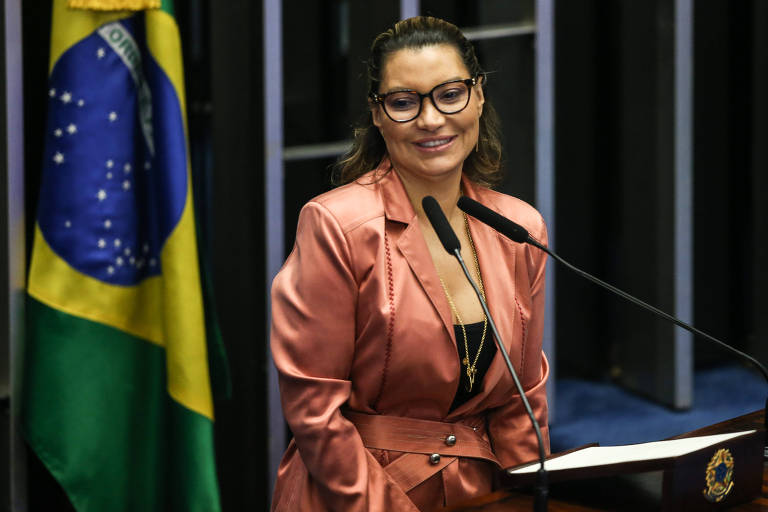 Uma mulher está discursando em um púlpito. Ela usa óculos e veste um blazer rosa brilhante sobre uma blusa preta. Ao fundo, há uma bandeira do Brasil.