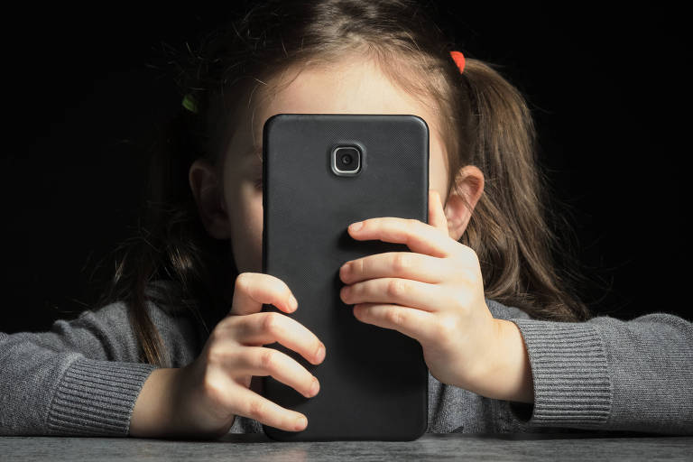 Fotografia colorida mostra close de criança vendo tela de celular na altura do rosto