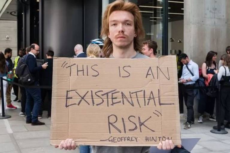 Imagem mostra homem segurando cartaz de papelão com os dizeres "This is an existential risk" ("Isso é uma crise existencial") em meio a uma praça, onde há outras pessoas.