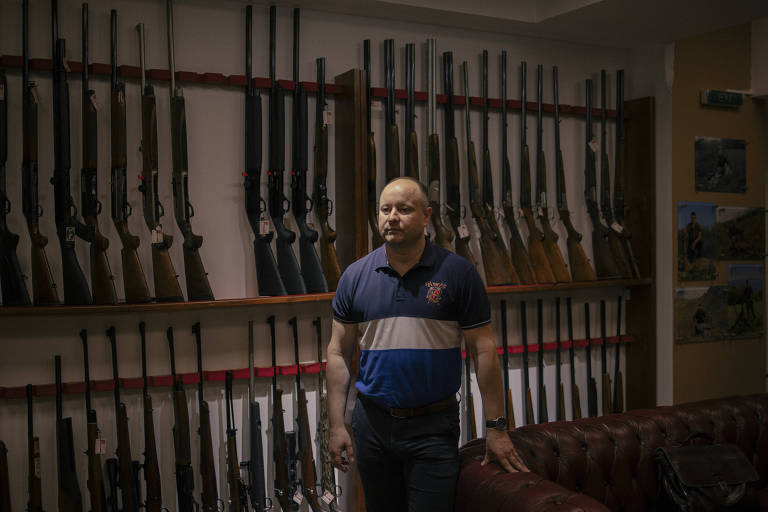 Homem careca com armas expostas em parede ao fundo