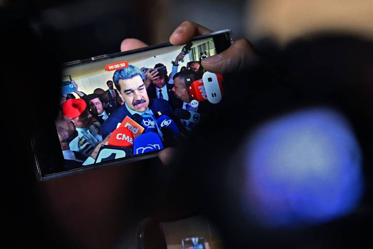 Um homem moreno, de bigode, usando terno e gravata, está cercado por microfones de jornalistas; a imagem aparece na tela de um celular que grava a cena