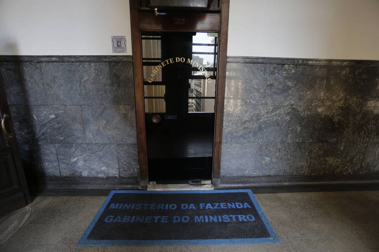 Na imagem se vê parte de um corredor, onde está uma porta de vidro na qual está escrito 'gabinete do ministro', no tapete à sua frente lê-se "ministério da fazenda, gabinete do ministro"