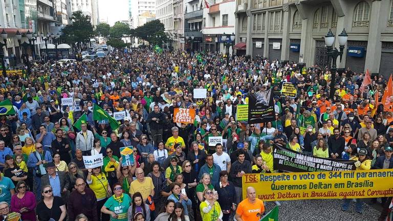 Multidão se reúne na extensão de aproximadamente uma quadra. Várias delas vestem camisetas amarelas ou verdes. Também há bandeiras do Brasil e uma faixa que diz "TSE respeite o voto de 344 mil paranaenses"