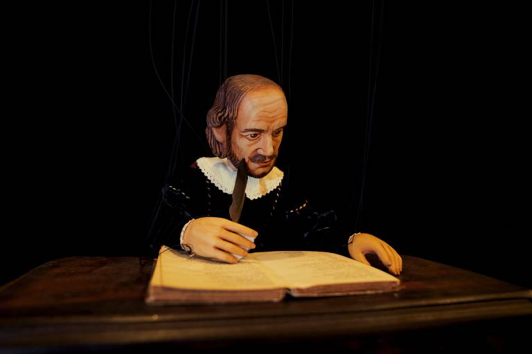 Um boneco homem com trajes do século 16 está sentando e com um caderno à sua frente