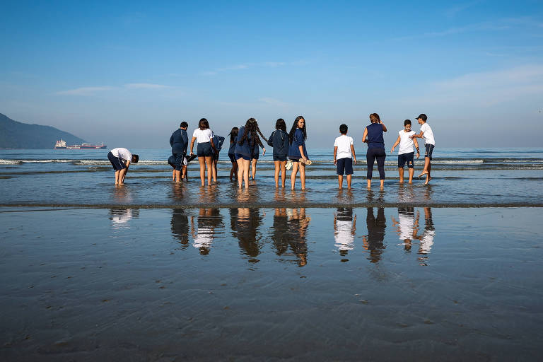 Imagem colorida mostra um grupo de crianças, com uniforme escolar, na praia, entre a areia e o mar.
