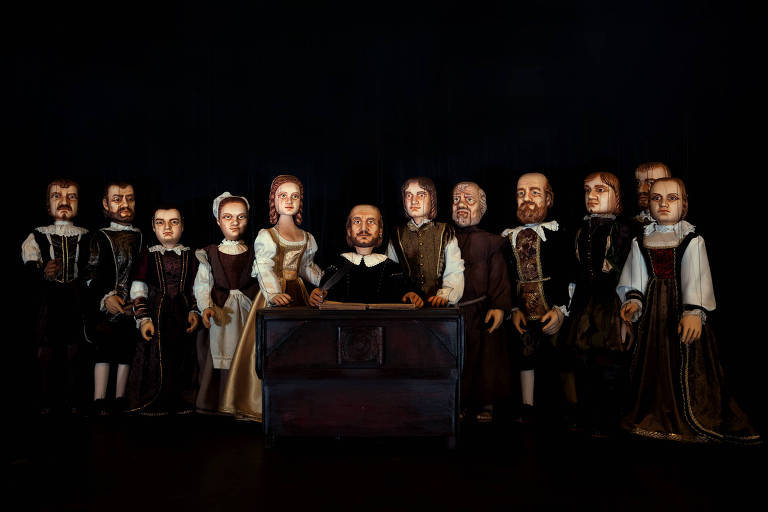 12 marionetes com trajes típicos do século 16 estão dispostas lado a lado num palco escuro
