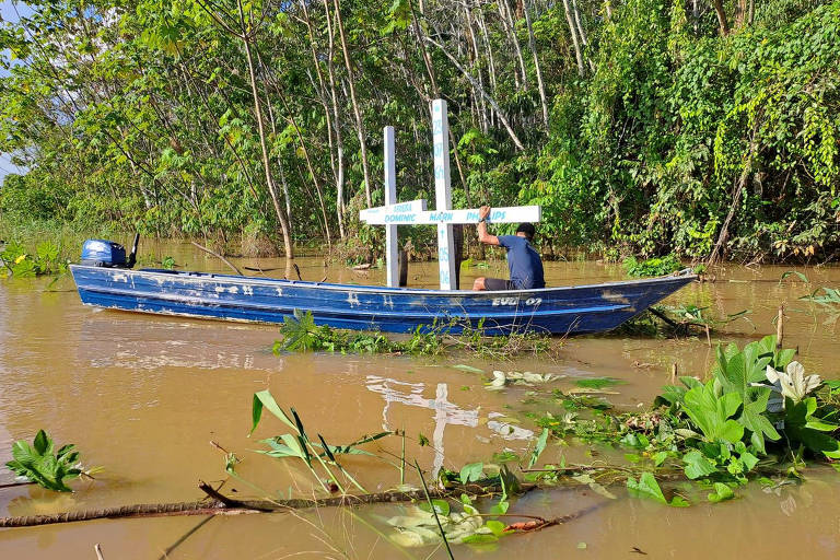 Em barco, homem coloca duas cruzes brancas com escritos em azul dentro de rio