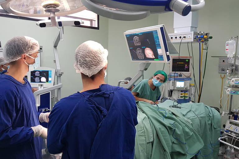 A imagem mostra uma sala de cirurgia com três profissionais de saúde. Dois deles， de costas， estão usando uniformes azuis e toucas cirúrgicas. Um terceiro profissional， visível à frente， está vestido com um avental verde e parece estar realizando um procedimento cirúrgico em um paciente coberto por um lençol verde. Ao fundo， há equipamentos médicos e um monitor exibindo informações.
