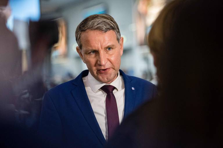 O político alemão Björn Höcke é um homem branco e loiro, vestido de terno azul e gravata vinho. Ele fala com um jornalista que está fora do enquadramento da foto 