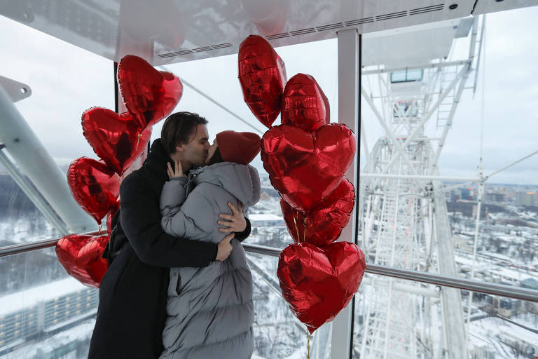 Homem vestido com casado preto beija mulher vestida com casado cinza-claro e gorro bonina. Eles estão circundados por balões em forma de coração vermelho, e dentro de uma roda gigante branca.