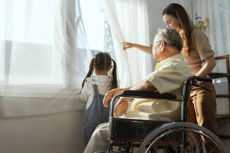 Mulher em frente a uma janela com um idoso em uma cadeira de rodas e uma menina em pé ao lado