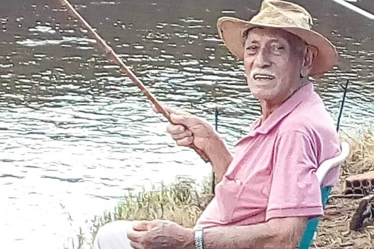 O comerciante Moisés Pereira à beira de um rio, sentado em uma cadeira de praia, segurando uma vara de pesca na mão direita
