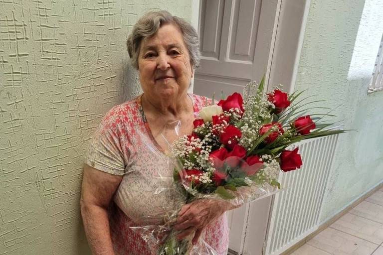 Maura Maria da Silva de pé com a ajuda de bengala segurando um buquê de flores vermelhas na outra mão