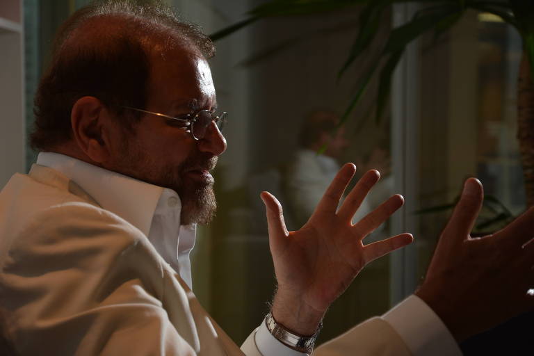 Retrato do fundador da CVC, Guilherme Paulus, de perfil, na penumbra, gesticulando enquanto fala. Vestido com uma camisa branca, ele usa óculos e relógio.