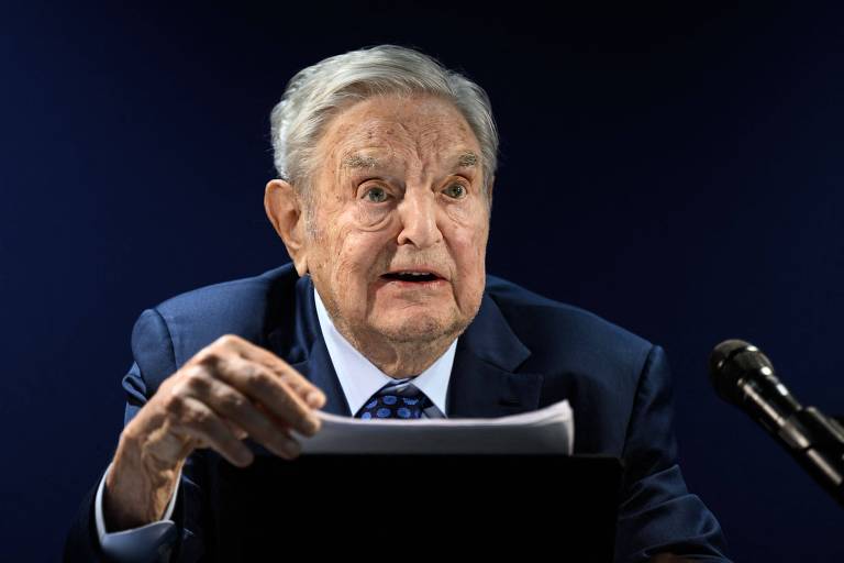 A imagem colorida mostra George Soros. Ele é um homem branco idoso, de cabelos brancos, veste terno e fala durante uma coletiva de imprensa