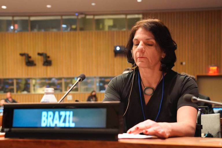Secretária está sentada à mesa, com uma roupa escura, olhando para baixo. À sua frente, uma placa de identificação escrito "Brazil", com a grafia em inglês