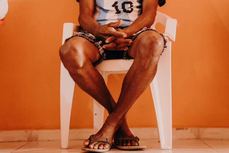 Homem está sentado em uma cadeira de plástico branca com as pernas cruzadas na altura do tornozelo e mãos encaixadas. Vemos ele da cintura para baixo, usando uma camisa branca com número 10 escrito e short colorido.