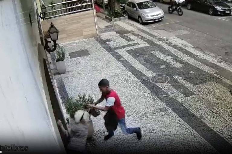 Homem de camisa vermelha e calça azul puxa bolsa de mulher idosa, que havia acabado de entrar na portaria de um prédio. Roubo aconteceu no bairro do Flamengo e foi registrado por câmera de segurança.