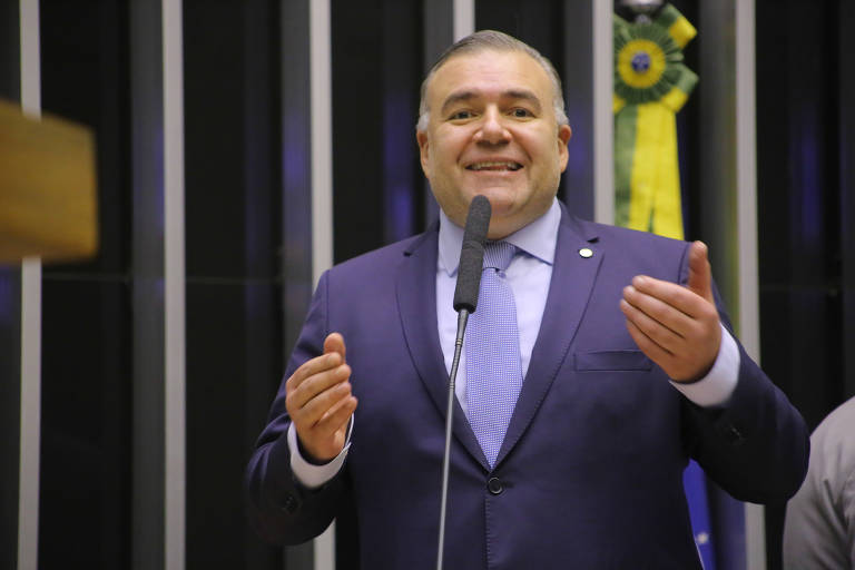 A imagem mostra Ney Leprevost, um homem de terno azul e gravata azul claro, falando ao microfone. Ele está gesticulando com as mãos e parece estar em um ambiente formal, possivelmente uma sessão parlamentar. Ao fundo, há uma faixa verde e amarela com um emblema, sugerindo um contexto oficial brasileiro.