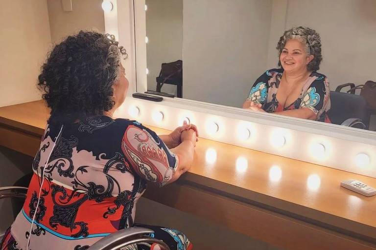 Mulher se olha em um espelho de camarim rodeado por luzes. Ela está sentada, tem cabelo cacheado com alguns fios brancos e usa um vestido colorido.
