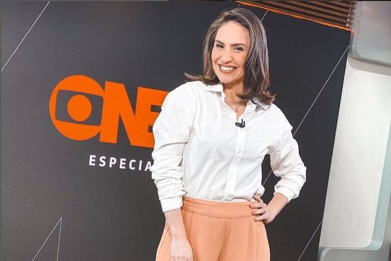 Em foto colorida, mulher de blusa branca posa a frente de uma logomarca da GloboNews