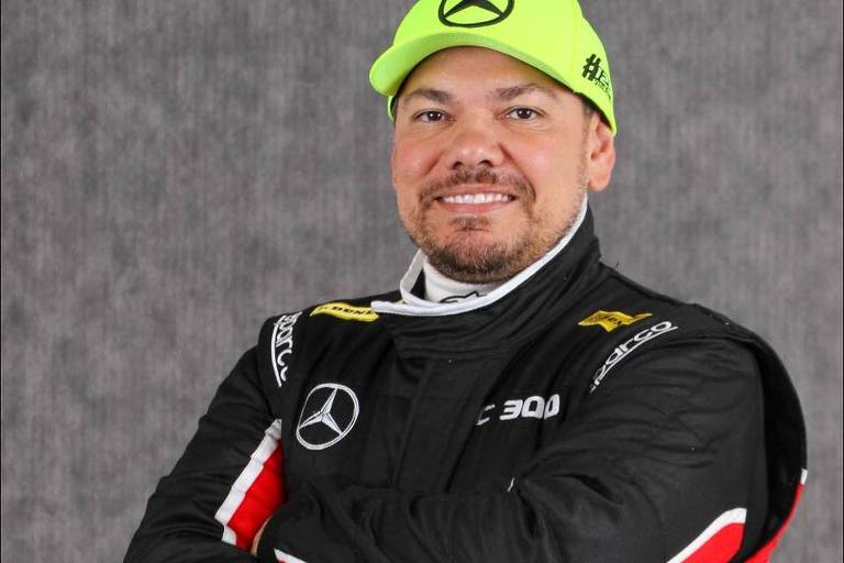 Douglas Pereira Costa, 42, de Jundiaí (SP), de braços cruzados; ele veste uniforme de piloto de corridas preto e vermelho e usa um boné verde com símbolo da Mercedes-Benz