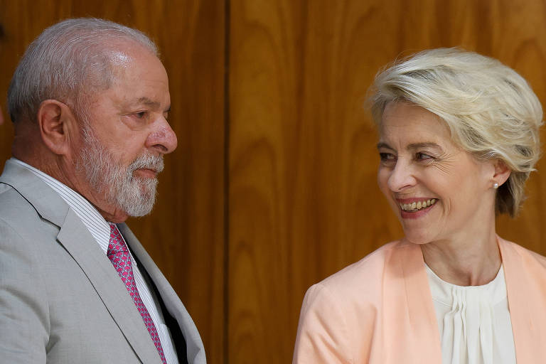 Lula de terno cinza claro olha pra direita e mulher de cabelos loiros/brancos de blusa branca e paletó rosa olha para ele e sorri