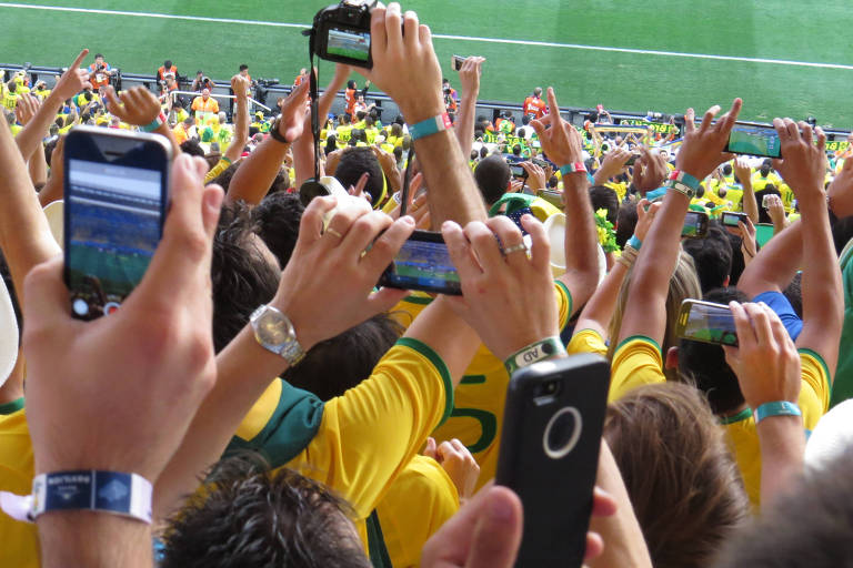 Torcedores uniformizados com a camisa do Brasil estão em estádio com seus celulares apontados para o campo, tirando foto do jogo do Brasil contra a Croácia na copa do mundo de futebol de 2014.