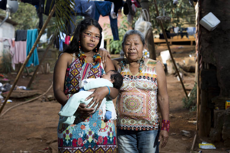 Duas mulheres indígenas olham para a câmera, uma delas jovem com pintura abaixo dos olhos, que segura um bebê de roupa azul, e outra com cabelos brancos, em um cenário com terra batida ao fundo, varais com roupas penduradas e mata