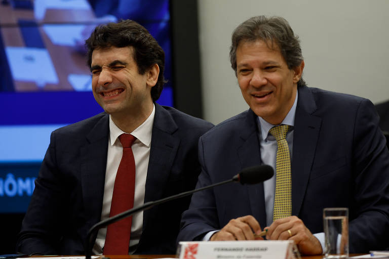 O ministro da Fazenda, Fernando Haddad (dir.), e Gabriel Galípolo; eles sorriem, são homens brancos, de cabelos lisos e castanhos, usam ternos escuros, camisas brancas e gravatas amarela e vermelha, respectivamente