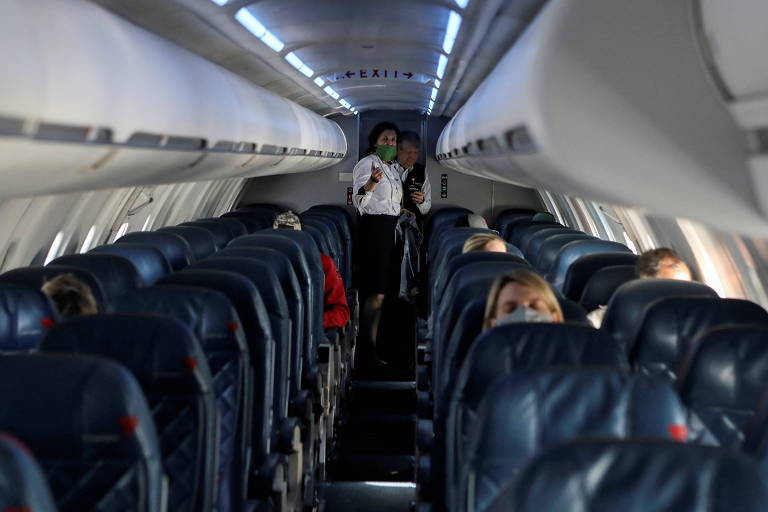 Vê-se um corrredor no meio dos assentos de uma aeronave, com uma funcionária da companhia aérea ao fundo