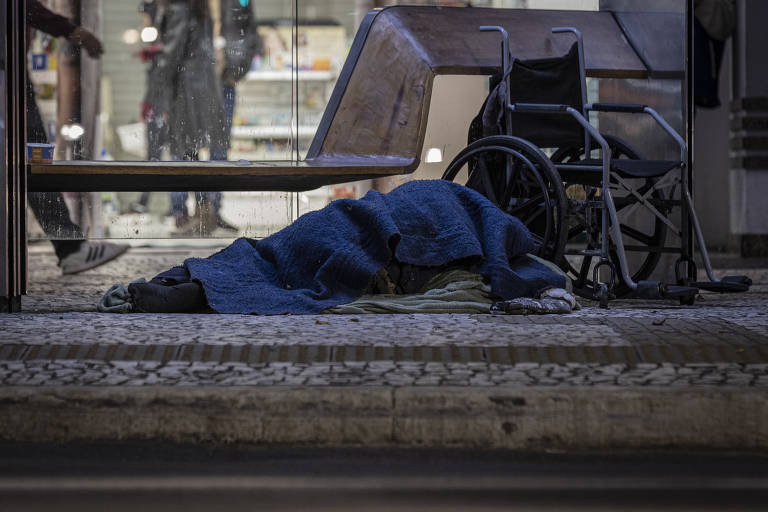 Imagem mostra uma pessoa enrolada num cobertor azul, deitada ao lado de uma cadeira de rodas, à noite, num ponto de ônibus