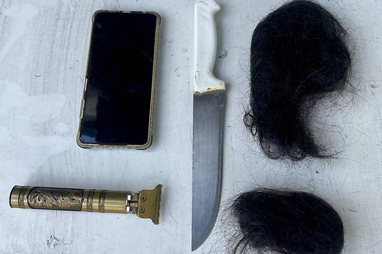 Celular, faca, máquina de cortar cabelo e duas mechas de cabelo dispostos em uma mesa branca