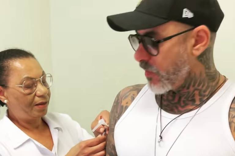 Em foto colorida, homem de camisa branca e todo tatuado recebe vacina em um posto de saúde