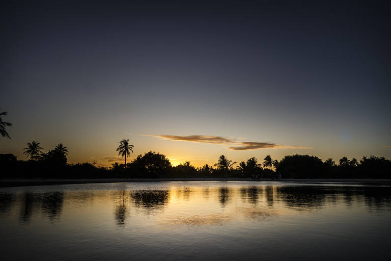 Em primeiro plano, há um lago que reflete a paisagem do pôr do sol junto à vegetação de coqueiros ao fundo