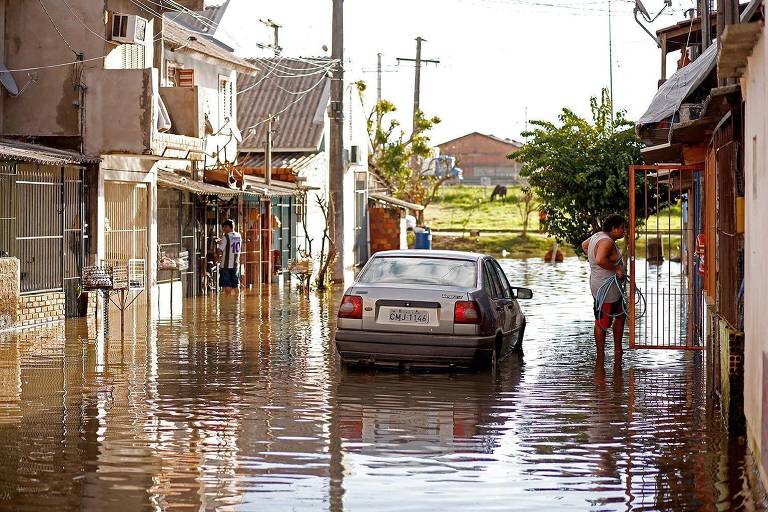 Rua alagada com pessoas com água pela canela e um carro com pneus cobertos de água