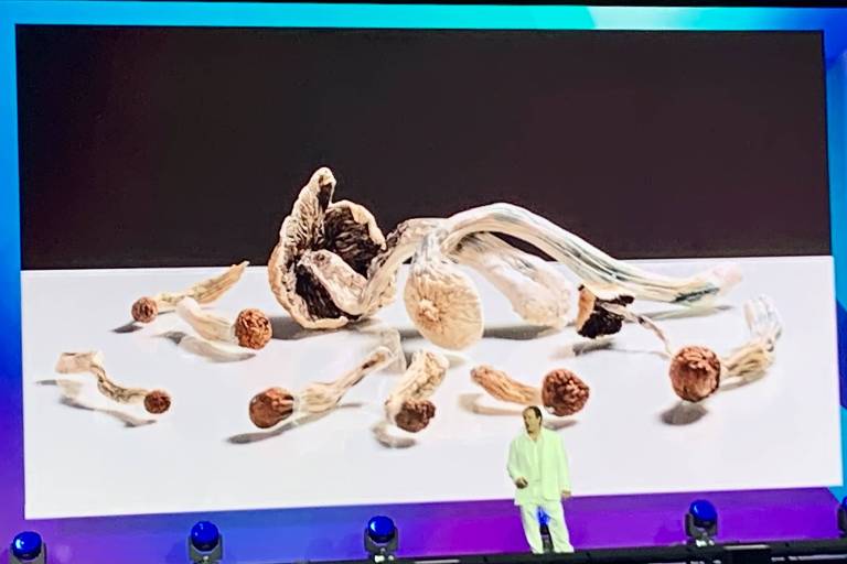 Homem de branco fala em conferência com telão mostrando imagens de cogumelos