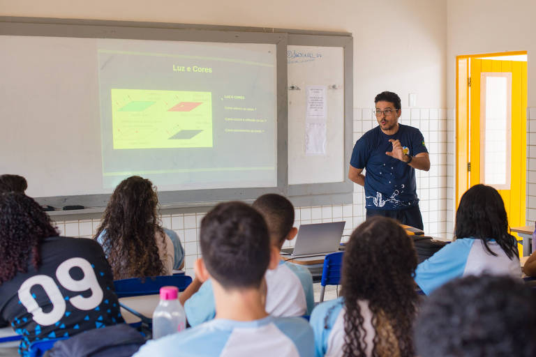 Imagem colorida mostra uma sala de aula, onde um homem está em pé, ao lado de um quadro branco. Ele conversa com alunos adolescentes que estão sentados em carteiras, de costas