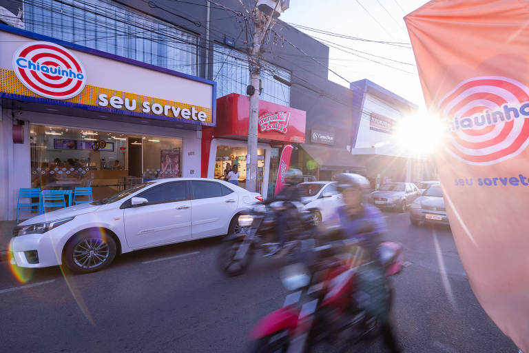 Fachada da loja da Chiquinho Sorvetes em destaque, ao lado de lojas do Mercadão do Óculos e da Cacau Show em rua no município de José Bonifácio (SP) onde há carros estacionados e motos passando