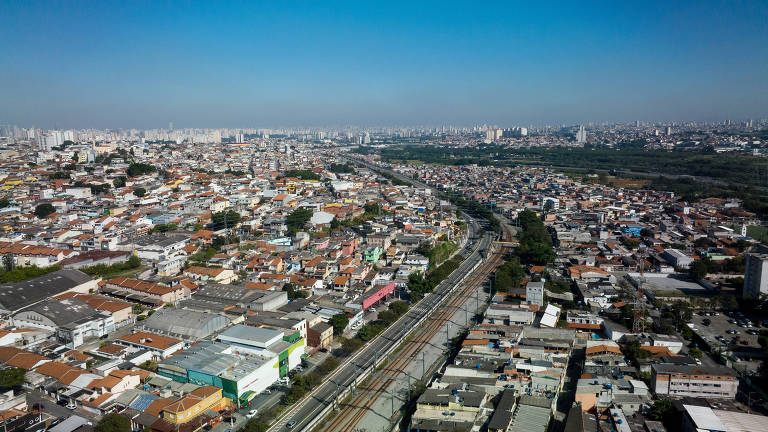 Vista aérea mostra avenida e ferrovia ao centro, cercadas por casas e prédios baicos de ambos os lados