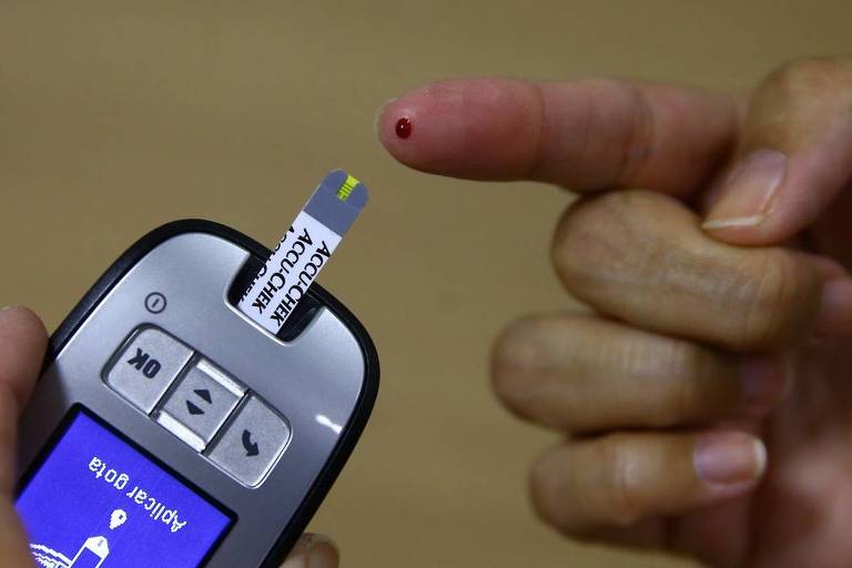 Mão branca com um pouco de sangue no dedo indicador e uma aparelho medidor de insulina ao lado. 