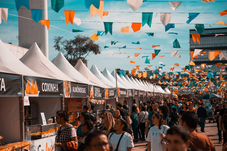 imagem captura festa popular, com barracas alinhadas sob um céu adornado por bandeirolas coloridas. Uma multidão de visitantes passeia entre as barracas