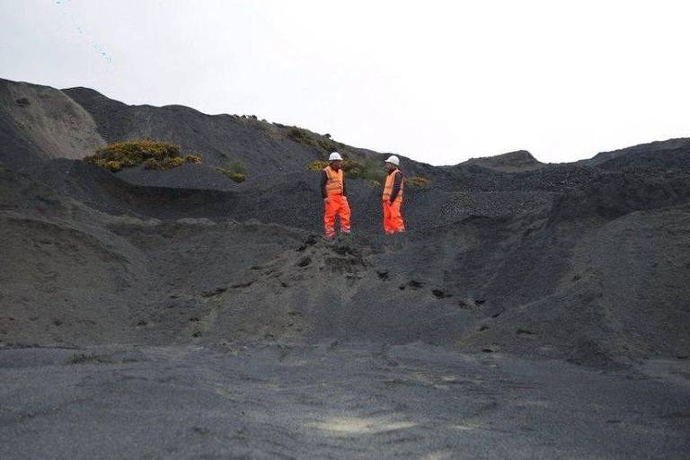 Dois homens de uniforme laranja conversam entre montes de rocha negra